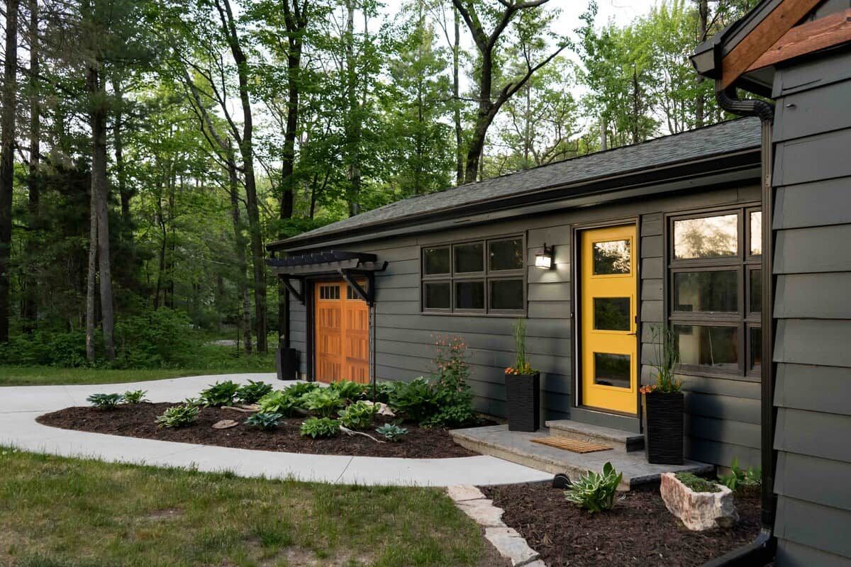 Une maison moderne à l’extérieur sombre, avec une porte d’entrée jaune et un jardin bien entretenu où poussent différentes espèces de plantes.