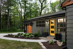 Une maison moderne à l’extérieur sombre, avec une porte d’entrée jaune et un jardin bien entretenu où poussent différentes espèces de plantes.
