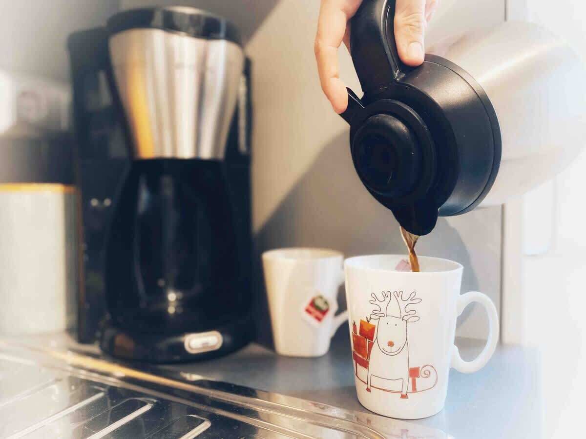 Une main verse du café d’une cafetière noire dans une tasse blanche ornée d’un motif de renne.
