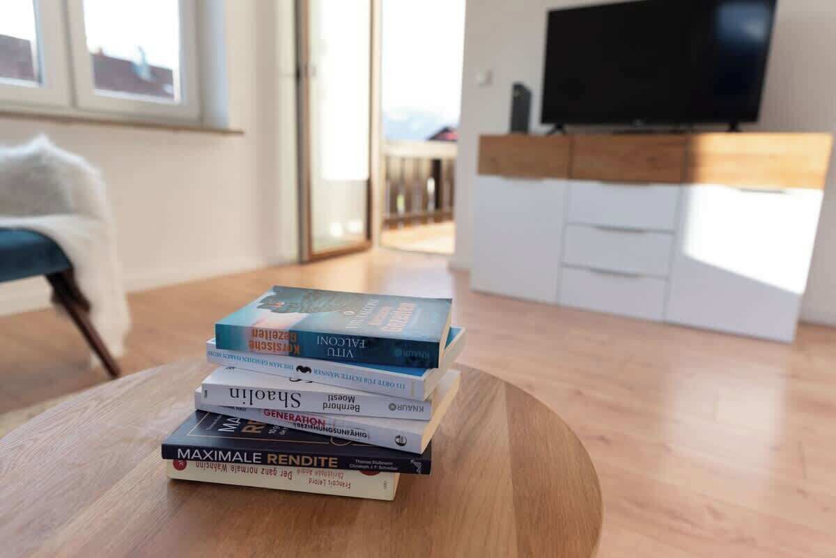 Ein Stapel Bücher auf einem Holztisch in einem Zimmer mit Fernseher an der Wand und Blick ins Freie durch ein Fenster.