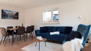 Una sala de estar moderna con un sofá azul, una mesa de comedor con sillas, una alfombra blanca y un cuadro en la pared.