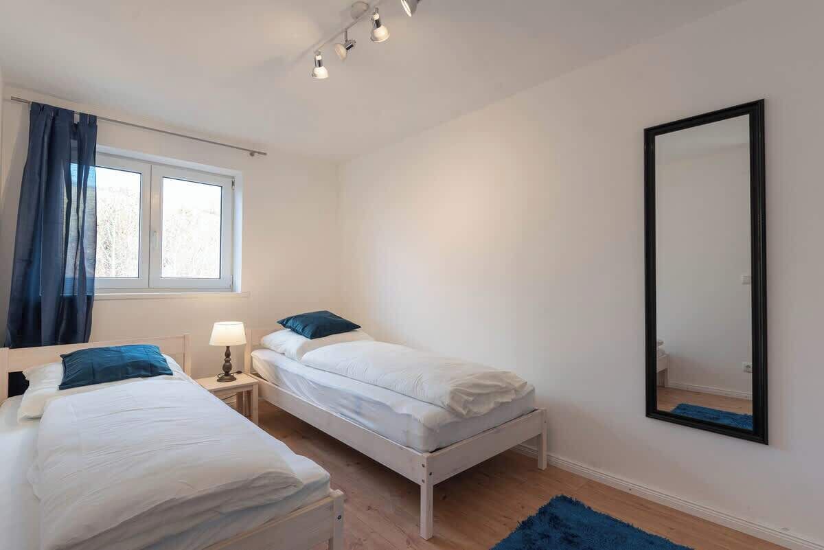 A bedroom with two beds, a window with curtains, a mirror, and a blue rug.