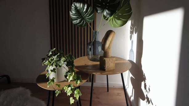 Una mesa redonda de madera con un jarrón y dos plantas verdes grandes, un jarrón blanco con plantas más pequeñas, y una escultura de madera.