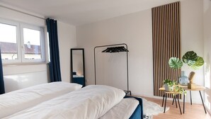 Un dormitorio con dos camas, una mesita de noche con plantas, una lámpara de pie y una ventana con cortinas.