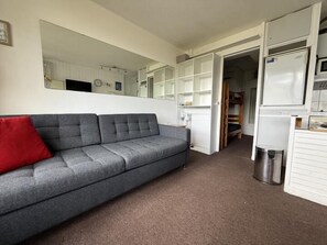 Un espacio habitable compacto con un sofá gris, un cojín rojo, un armario blanco y un espejo que refleja la habitación.