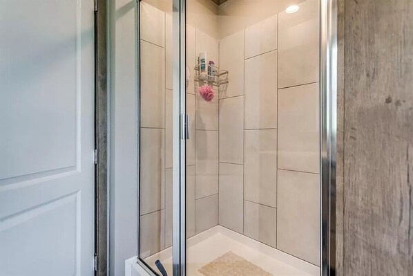 A shower with glass doors, beige tiles, and a small shelf with toiletries.