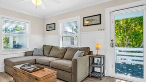A modern living room with a sectional sofa, a wooden coffee table, and a side table with a lamp. There are large windows with blinds, a door with a view of greenery, and framed artwork on the walls. -  Bungalow #3 - 2 Bed/1 Bath & Pool in Islamorada (Islamorada)