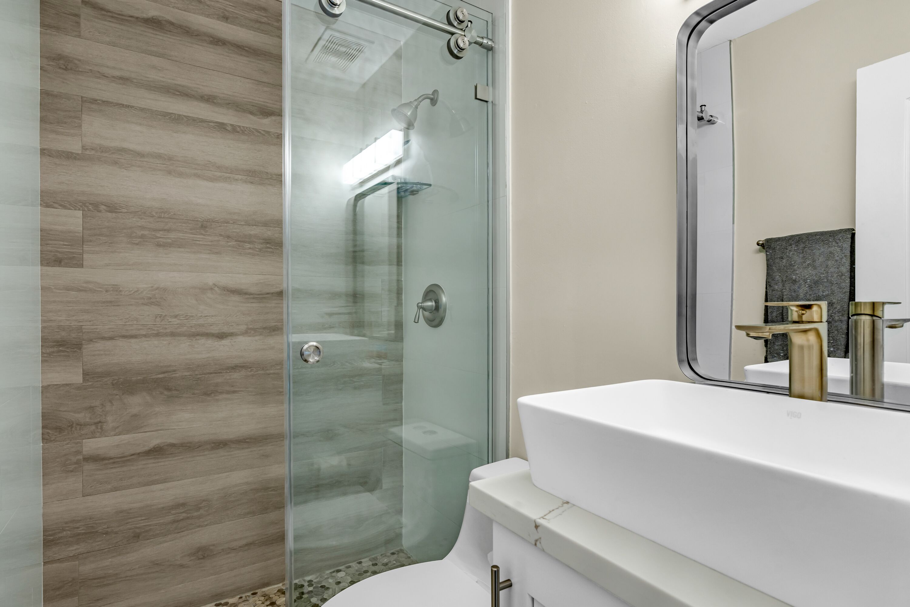 A modern bathroom with a glass-enclosed shower, a white sink with gold fixtures, and a mirror reflecting a toilet and sink.