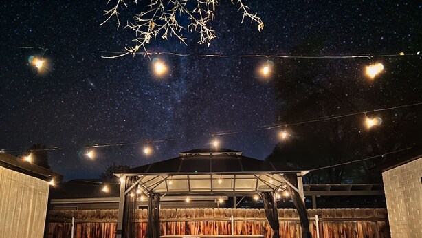 Un patio al aire libre con zona de estar cubierta, jacuzzi y luces colgantes bajo un cielo estrellado.