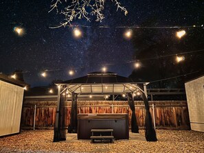 Ein Terrassenbereich im Freien mit überdachter Sitzecke, einem Whirlpool und Lichterketten vor einem sternenübersäten Nachthimmel.