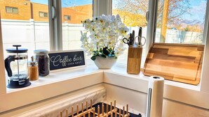 Una encimera de cocina con un letrero de cafetería, un molinillo de café, un tarro de azúcar, un florero y una tabla de cortar de madera.