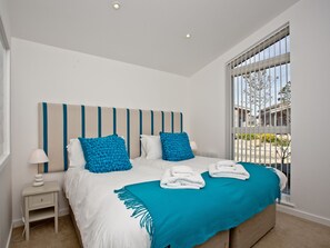 A bedroom with a bed, a nightstand, a window with blinds, and a view of the outside.