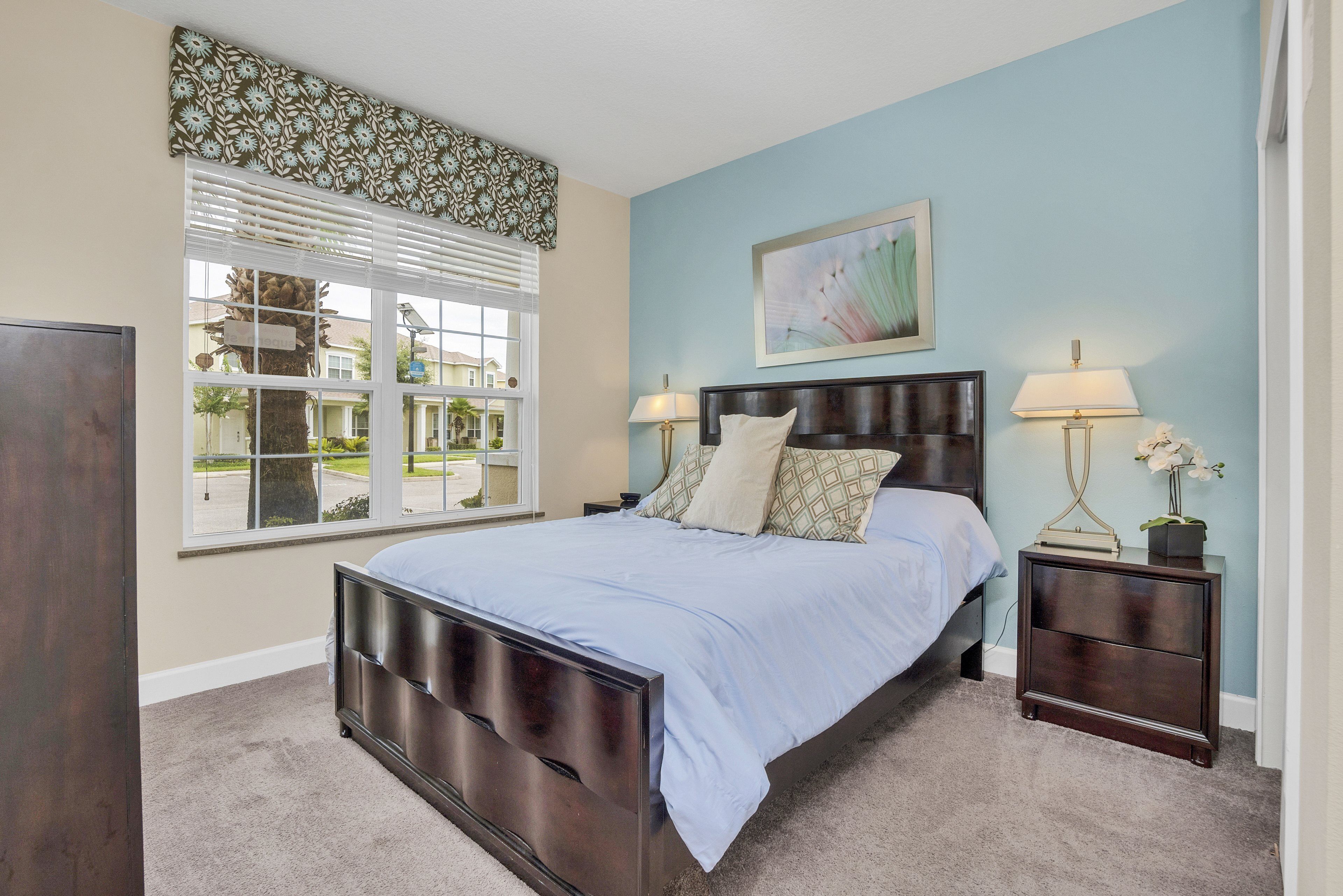 A bedroom with a large bed, two bedside tables with lamps, a window with blinds, and a wall-mounted artwork.