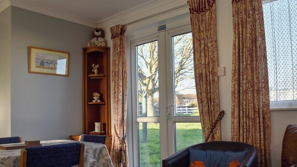 A dining area with a table set for two, a brown leather sofa, and a window with patterned curtains. - A wonderful chalet set in a great seaside location. (Kessingland)