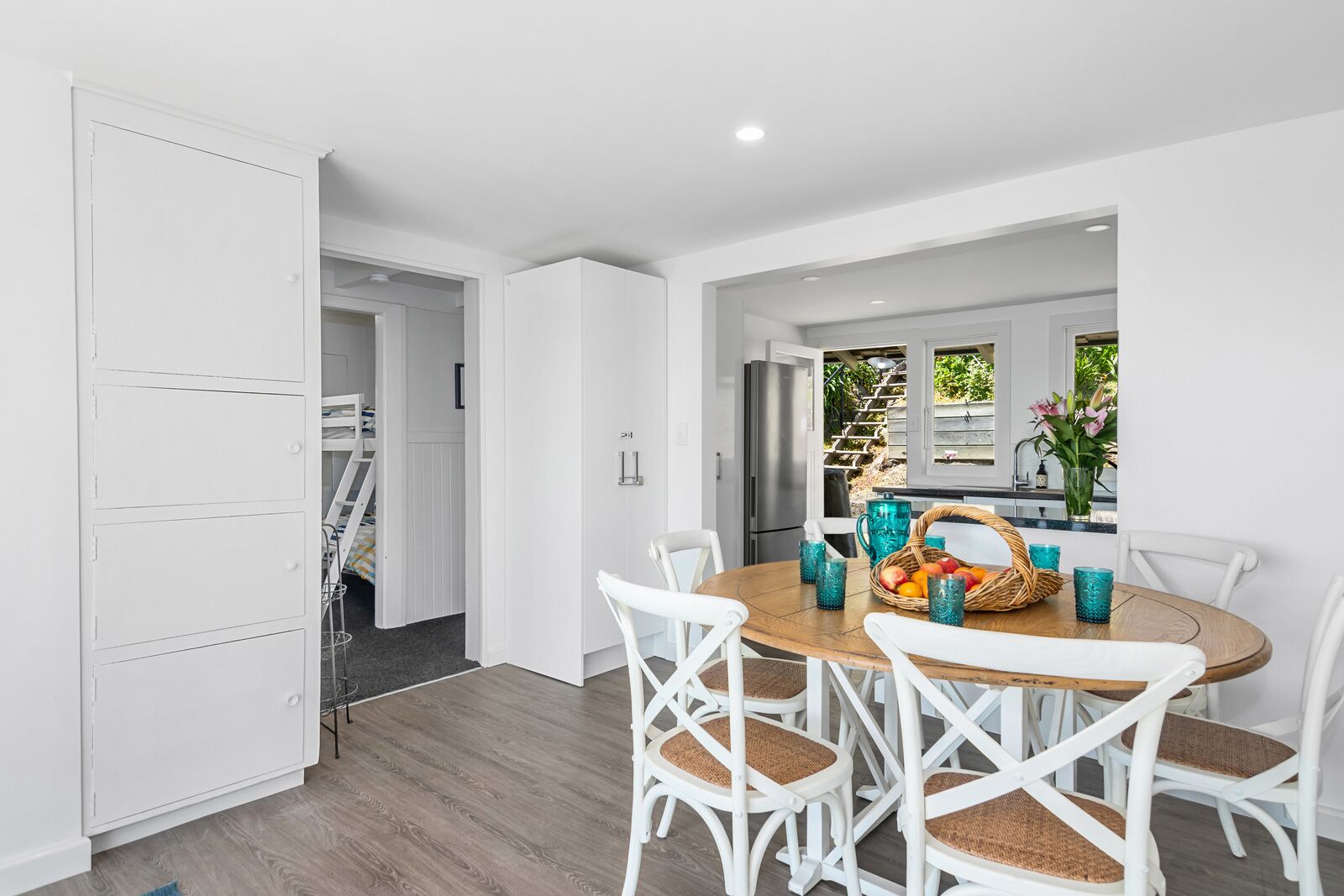 A modern kitchen with white cabinetry, a dining table with four chairs, and a view of a garden through a window.