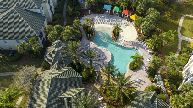Vista aérea de un complejo turístico con piscina, zonas de descanso y palmeras.