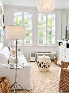 A well-lit living room with a white sofa, a floor lamp, a round ottoman, and a television on a white cabinet.