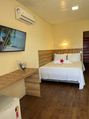 Quarto de hotel com cama, televisão, aparelho de ar condicionado e mesa de cabeceira de madeira.