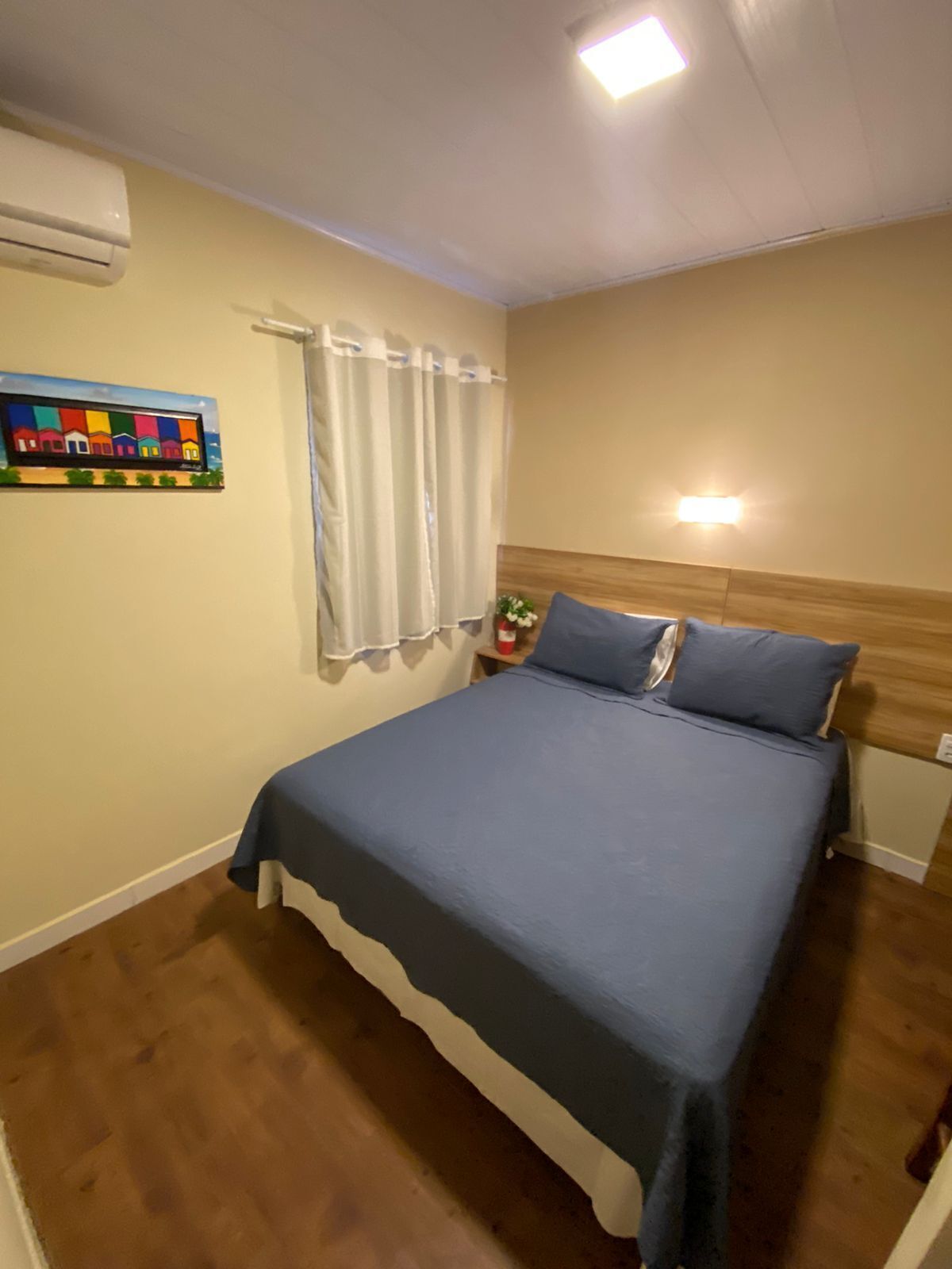 Un dormitorio con cama, cortinas, aire acondicionado instalado en la pared, mesita de noche con una planta y un cuadro en la pared.