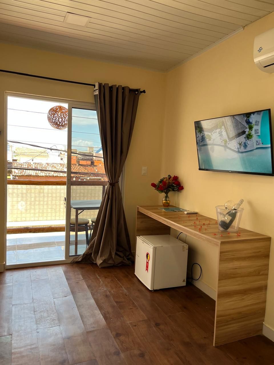 Una habitación con escritorio de madera, televisión, refrigerador pequeño, puerta corredera de vidrio y vista de edificios.