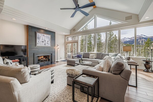 A spacious living room with a fireplace, large windows offering mountain views, and a ceiling fan.