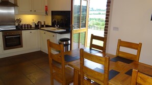 Een keuken met witte kasten, roestvrijstalen apparatuur, een houten eettafel met stoelen en uitzicht op groen door een raam.