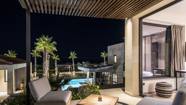 Un espace extérieur moderne aménagé avec des sièges, donnant sur la piscine, doté de mobilier en bois et d’une porte vitrée qui mène à un balcon.