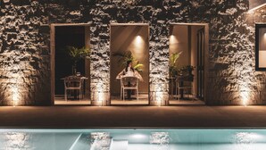 Un espace aménagé au bord de la piscine, avec des murs en pierre, trois portes donnant sur des coins salon et des plantes en pot.