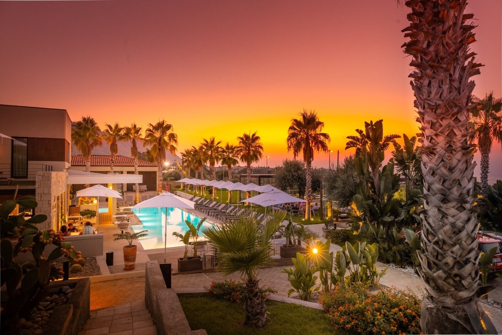 A resort with a swimming pool, lounge chairs, and palm trees against a sunset backdrop.