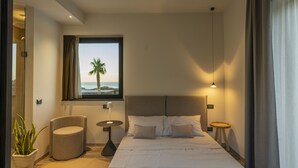 Une chambre d’hôtel avec un lit, un fauteuil rond, une petite table, une plante en pot et une fenêtre donnant sur la mer.