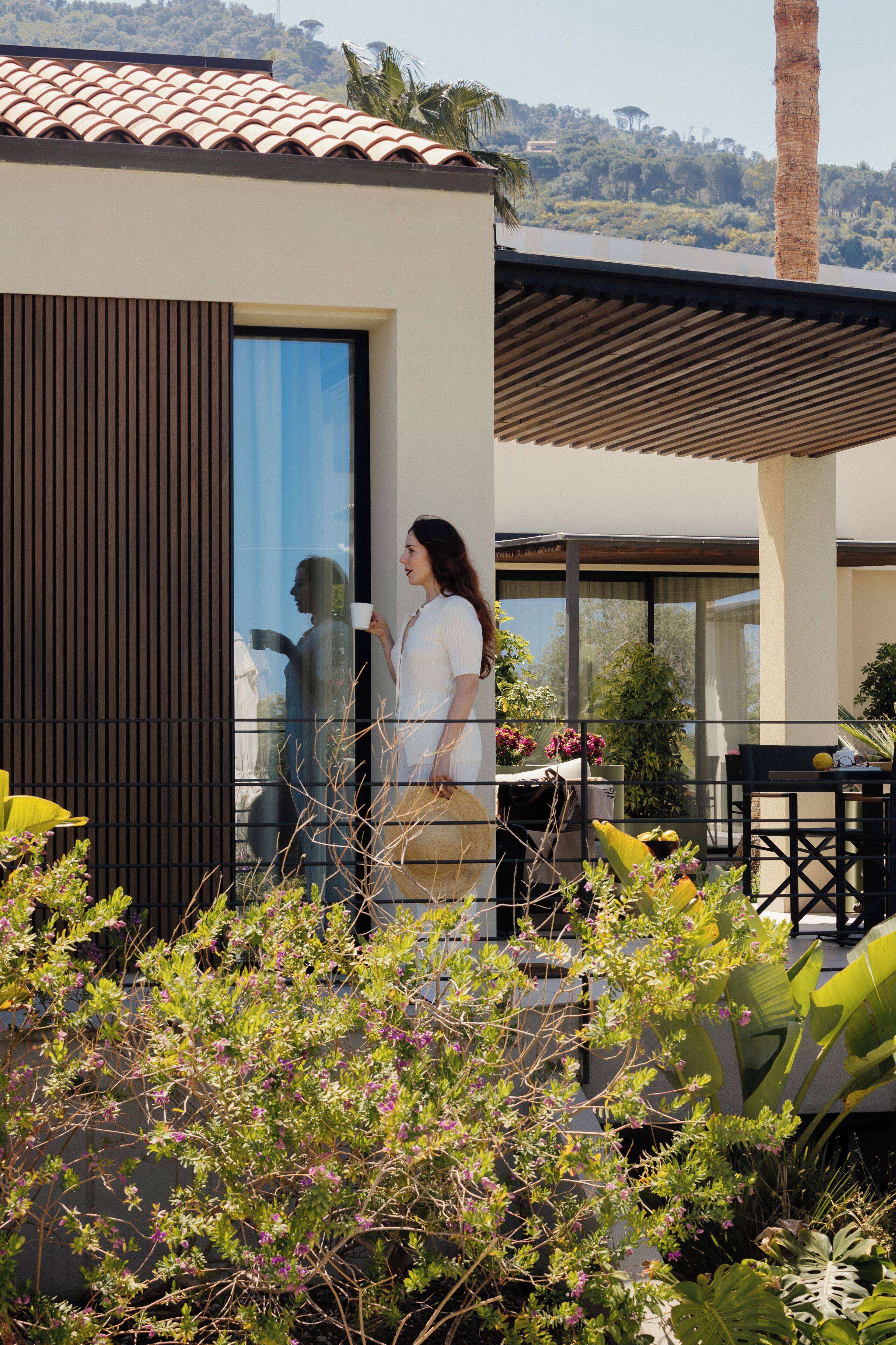 Une maison moderne dotée d’une grande porte vitrée, d’un patio agrémenté de plantes et offrant une vue sur une colline.