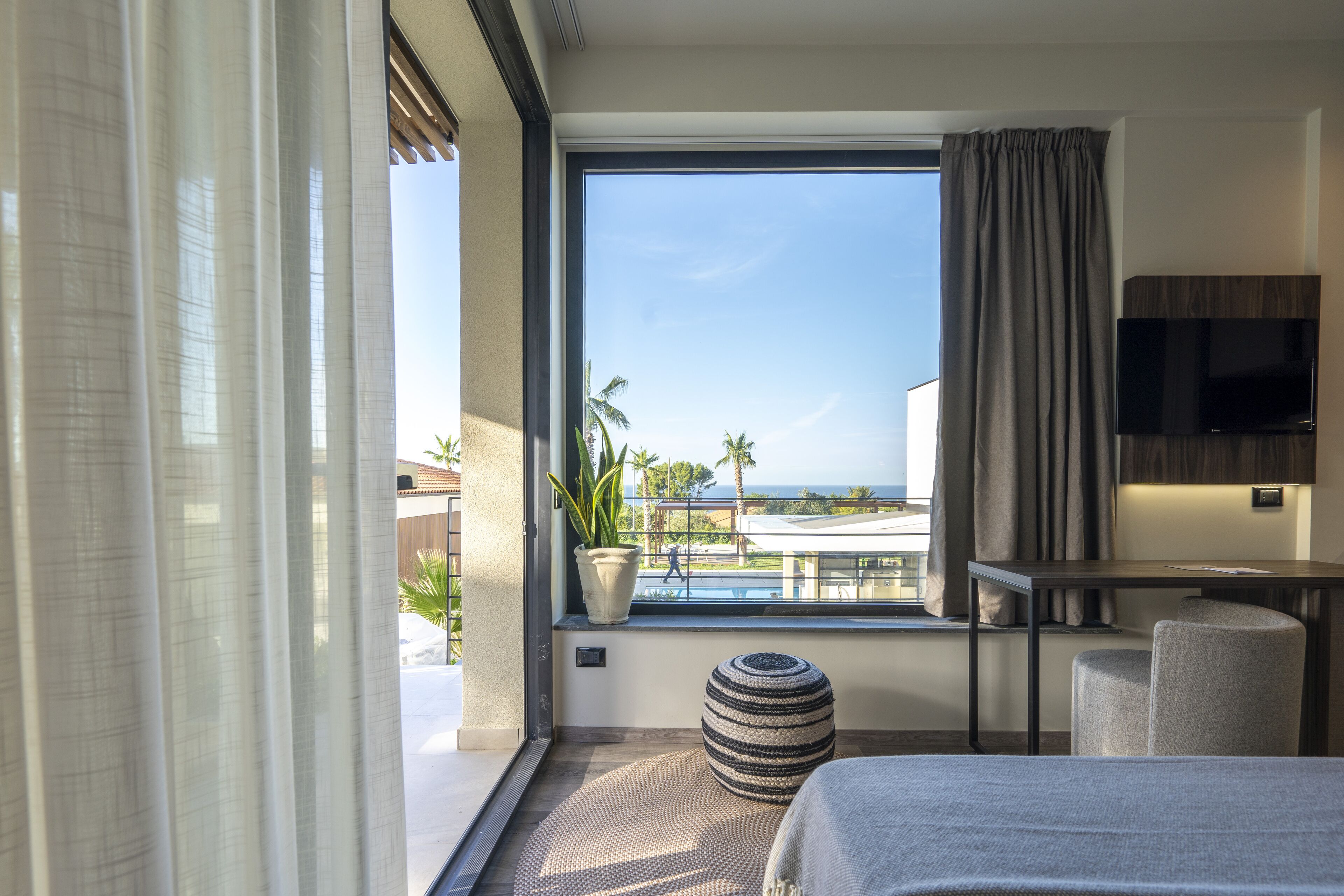 Ein modernes Hotelzimmer mit großem Fenster, das Blick auf Pool und Palmen bietet, einem flachbildfernseher an der Wand und einer gemütlichen Sitzecke mit Sofa und Hocker.