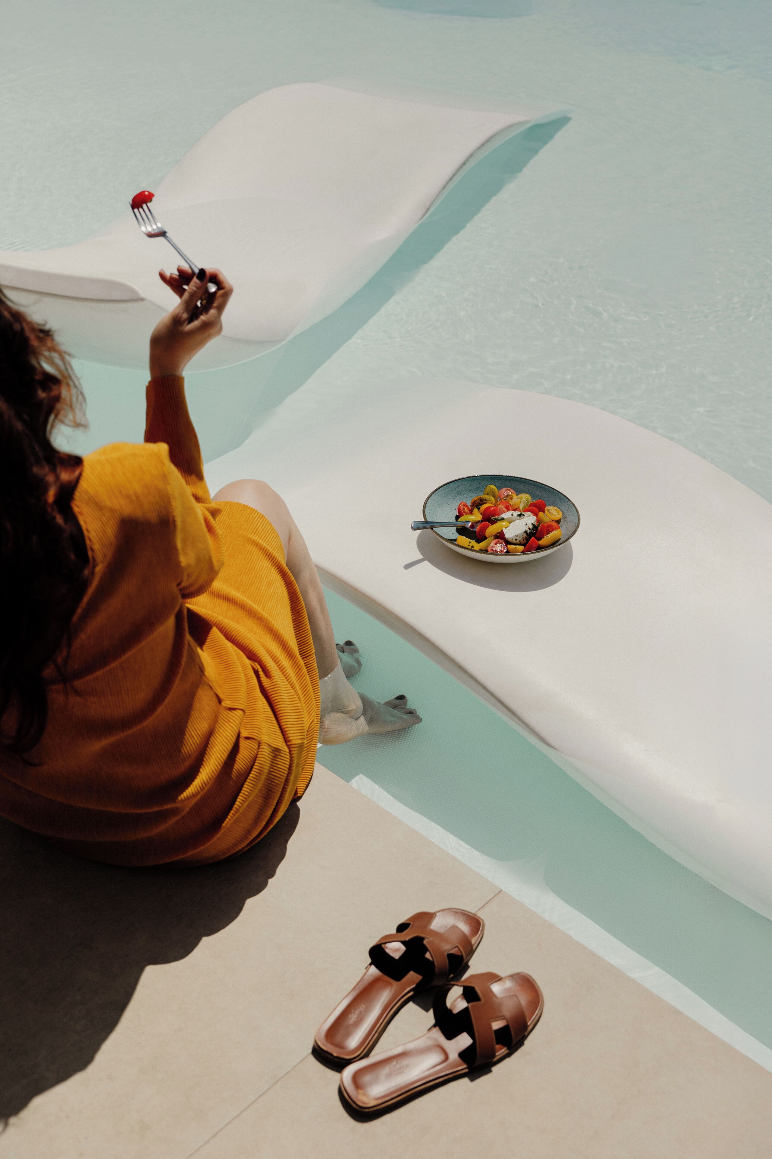 Una persona sentada junto a una piscina, con un cuenco de fruta y un par de sandalias.