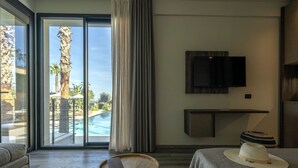 Une chambre d’hôtel dotée d’une grande porte-fenêtre coulissante donnant sur une piscine, d’un téléviseur à écran plat fixé au mur et d’un lit sur lequel est posé un chapeau.