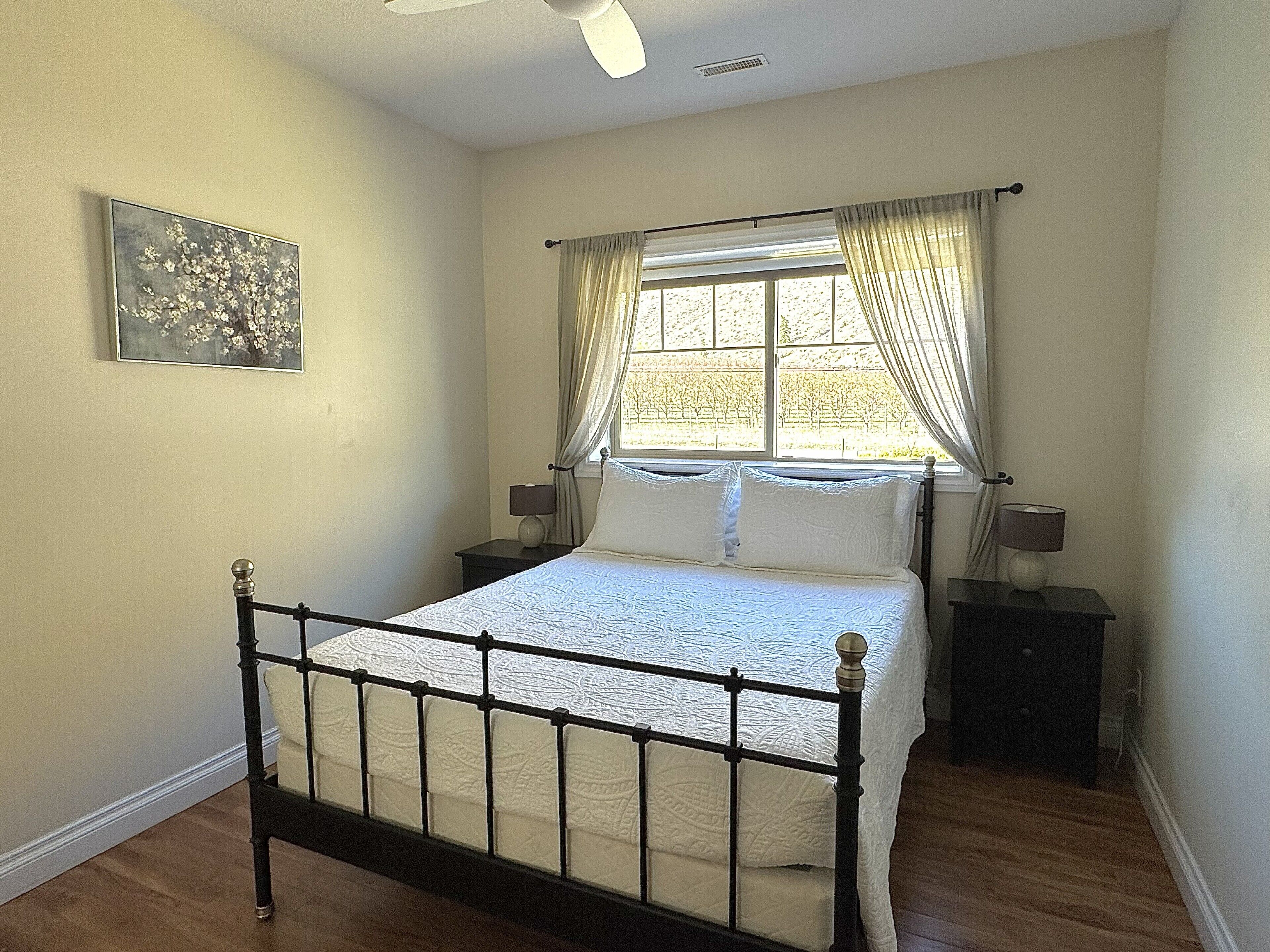 A bedroom with a bed, two bedside tables, a window with curtains, a ceiling fan, and a wall-mounted picture.