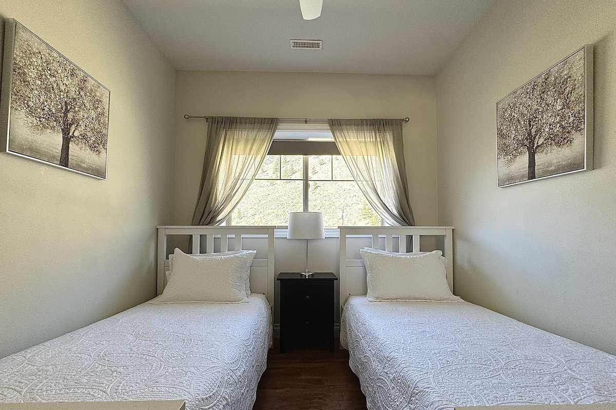 A bedroom with two single beds, a window with curtains, and two wall-mounted paintings.