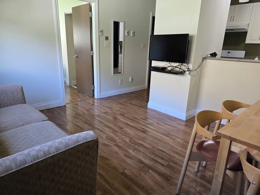 A living room with a sofa, a dining table with chairs, a TV mounted on the wall, and a kitchen area visible through an open door.