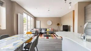 A modern kitchen with a central island, a dining area with a table and chairs, a sofa set, and a large window providing natural light. - Luxury London Garden Flat (London)