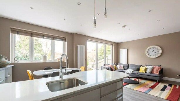 A modern kitchen with a central island, a dining area with a table and chairs, a grey sofa with colorful cushions, and a large window providing natural light. - Luxury London Garden Flat (London)