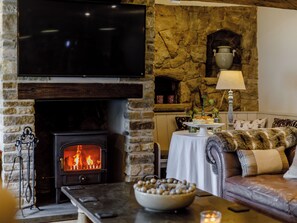 A cozy living room with a stone fireplace, a flat-screen TV, a leather sofa, and a dining table with a vase of flowers.