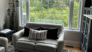 Ein Zimmer mit einem großen Fenster, einem grauen Sofa mit gemusterten Kissen, einer hohen Stehlampe und einer dunklen Vitrine.