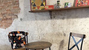 Twee stoelen met houten zitvlak en metalen frames, een klein rond tafeltje en een plank met gezelschapsspellen en boeken tegen een bakstenen muur.