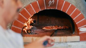 A person using a pizza peel to place a pizza into a brick oven. - Pitlane Lodge (Stavelot)
