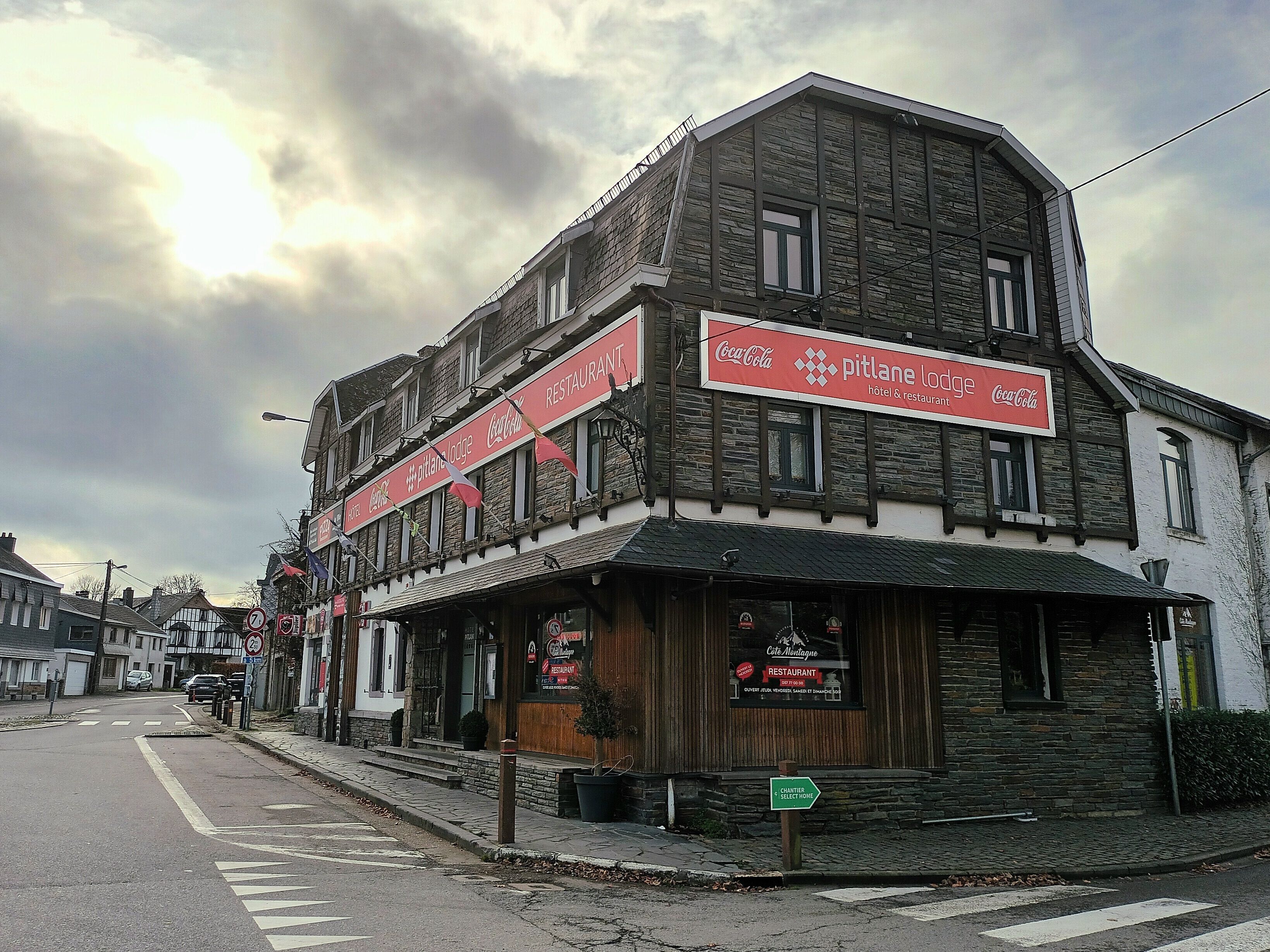 Ein zweistöckiges Gebäude mit einer Steinaußenfassade, einem Coca-Cola-Schild und einem Schild mit der Aufschrift 'Pitlane Lodge', das ein Hotel und Restaurant kennzeichnet.