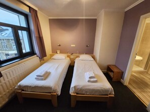 A hotel room with two single beds, a window with curtains, a radiator, a small wooden nightstand, and a shower area. - Pitlane Lodge (Stavelot)