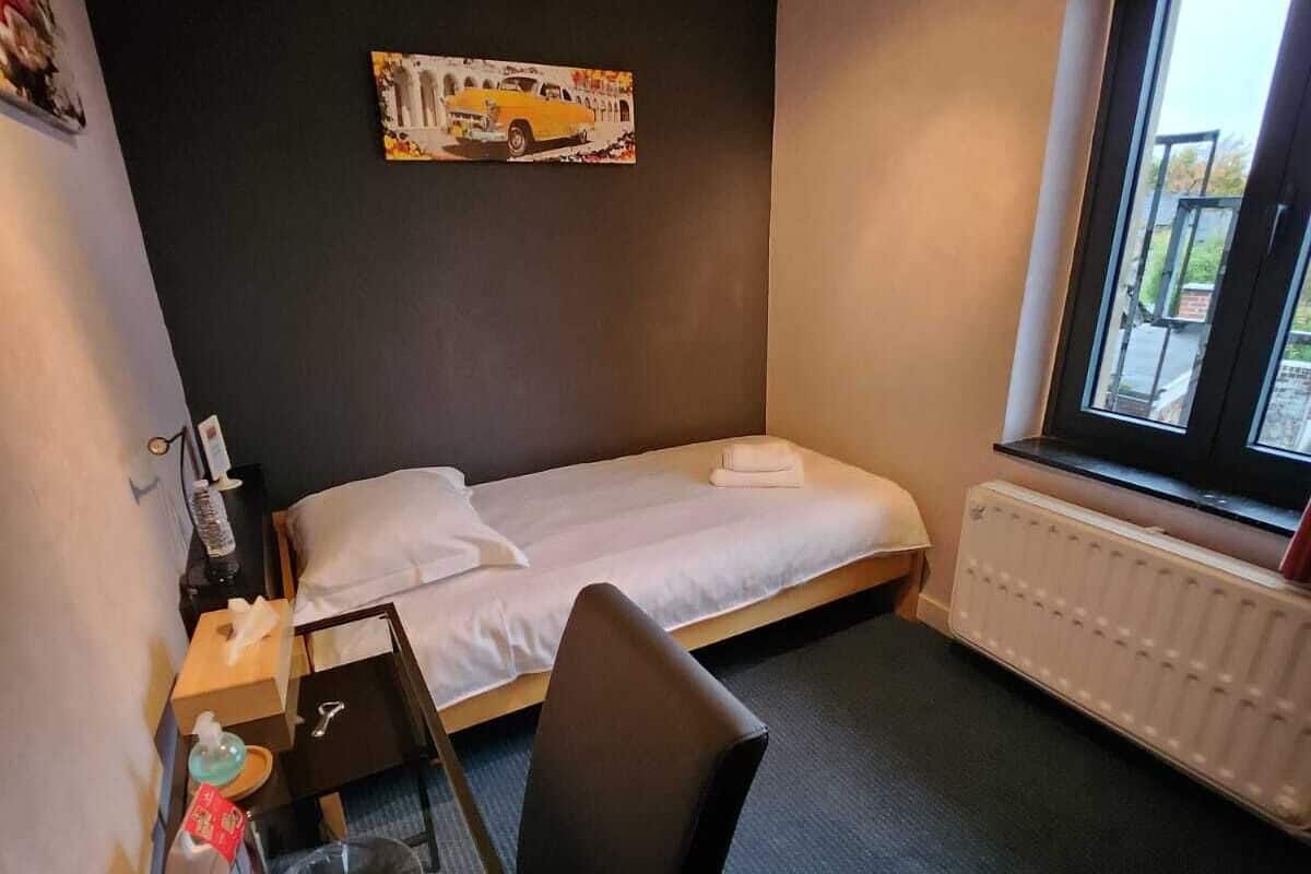 A hotel room with a single bed, a small table with a chair, a radiator, and a window with a view of the outside.
