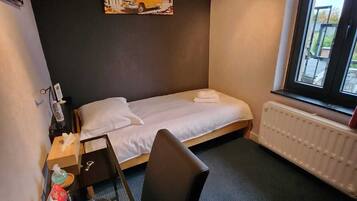 A hotel room with a single bed, a small table with a chair, a radiator, and a window with a view of the outside.