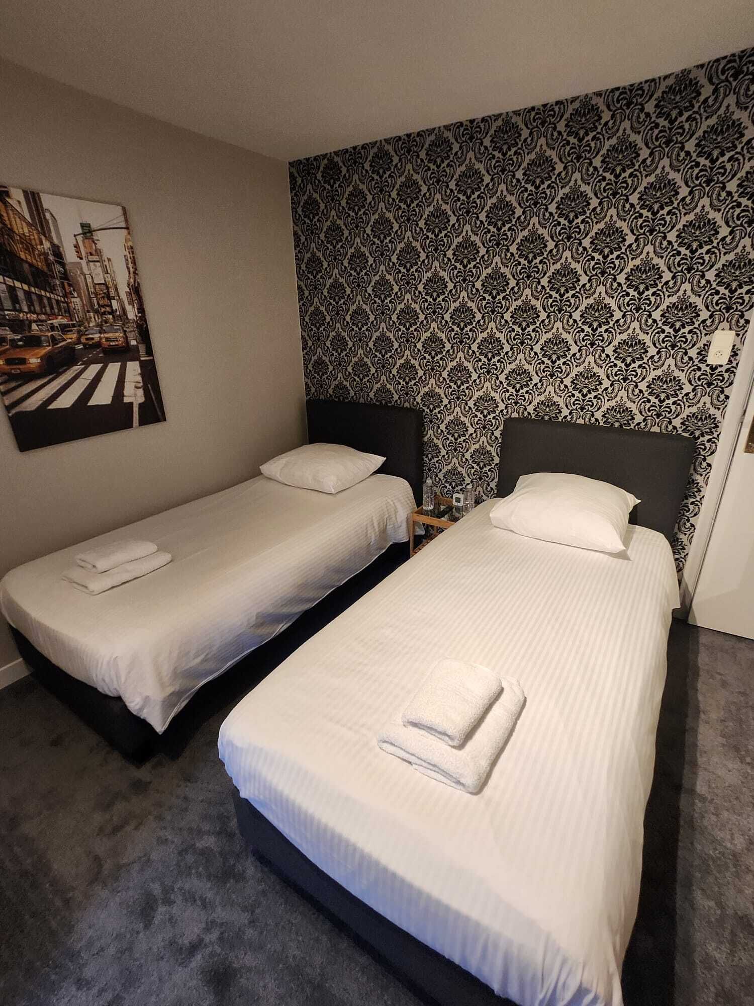 A hotel room with two single beds, a patterned wallpaper, and a cityscape painting on the wall.