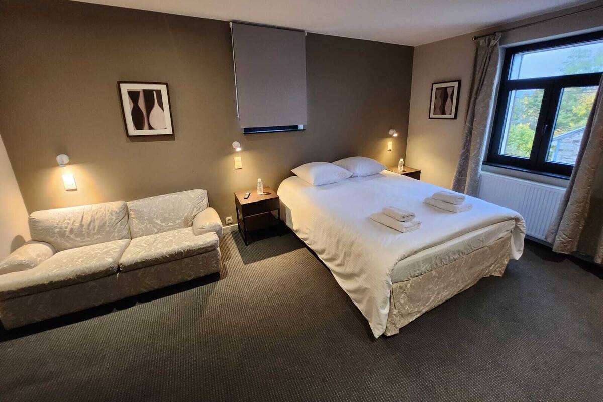 A hotel room with a bed, a sofa, two bedside tables, and a window with curtains.