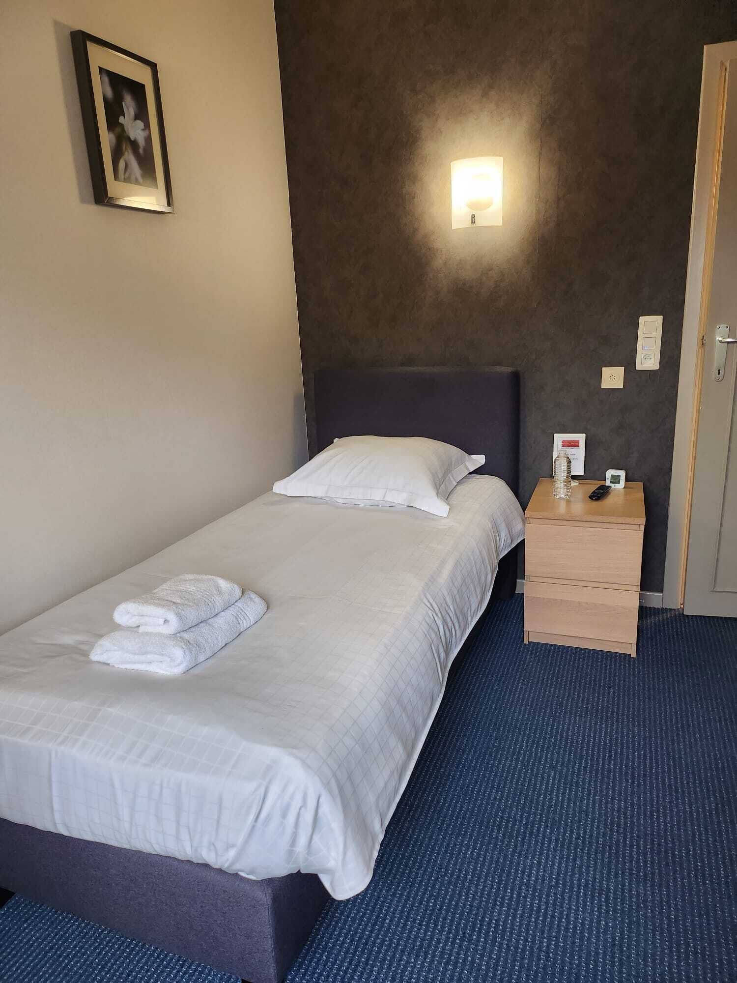 A neatly made bed with white linens and towels, a bedside table with a bottle and remote, and a wall-mounted light fixture.